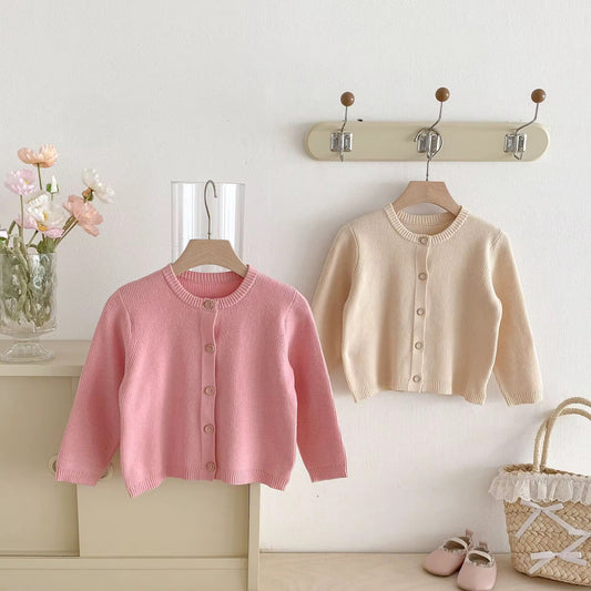 Two children's cardigans, one pink and one beige, hanging on a coat rack with a decorative basket and flowers in the background.